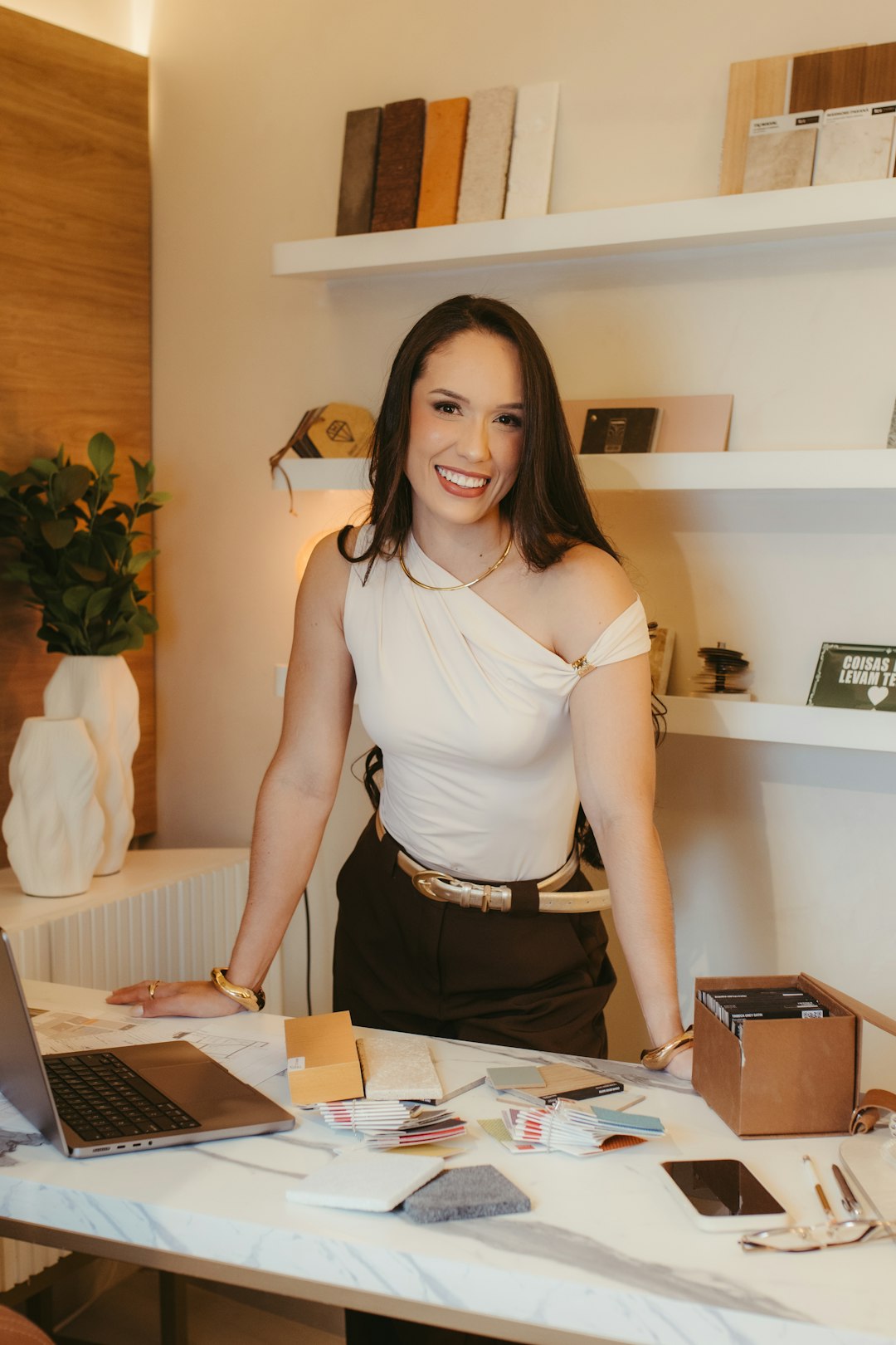 Emprendedora joven sonriendo en el estudio de su marca, con laptop y muestras de producto sobre el escritorio